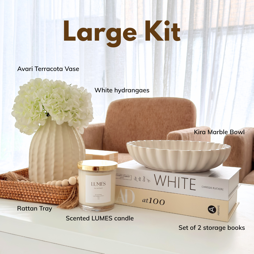A styled interior scene showing a curated home decor set on a white table in front of a sofa, with overlaid text labels naming the items: Avari Terracotta Vase, White hydrangeas, Kira Marble Bowl, Rattan Tray, LUMES candle, books, and a Large Kit title. Because a real room environment is visible, this is Lifestyle rather than Product/Packaging.