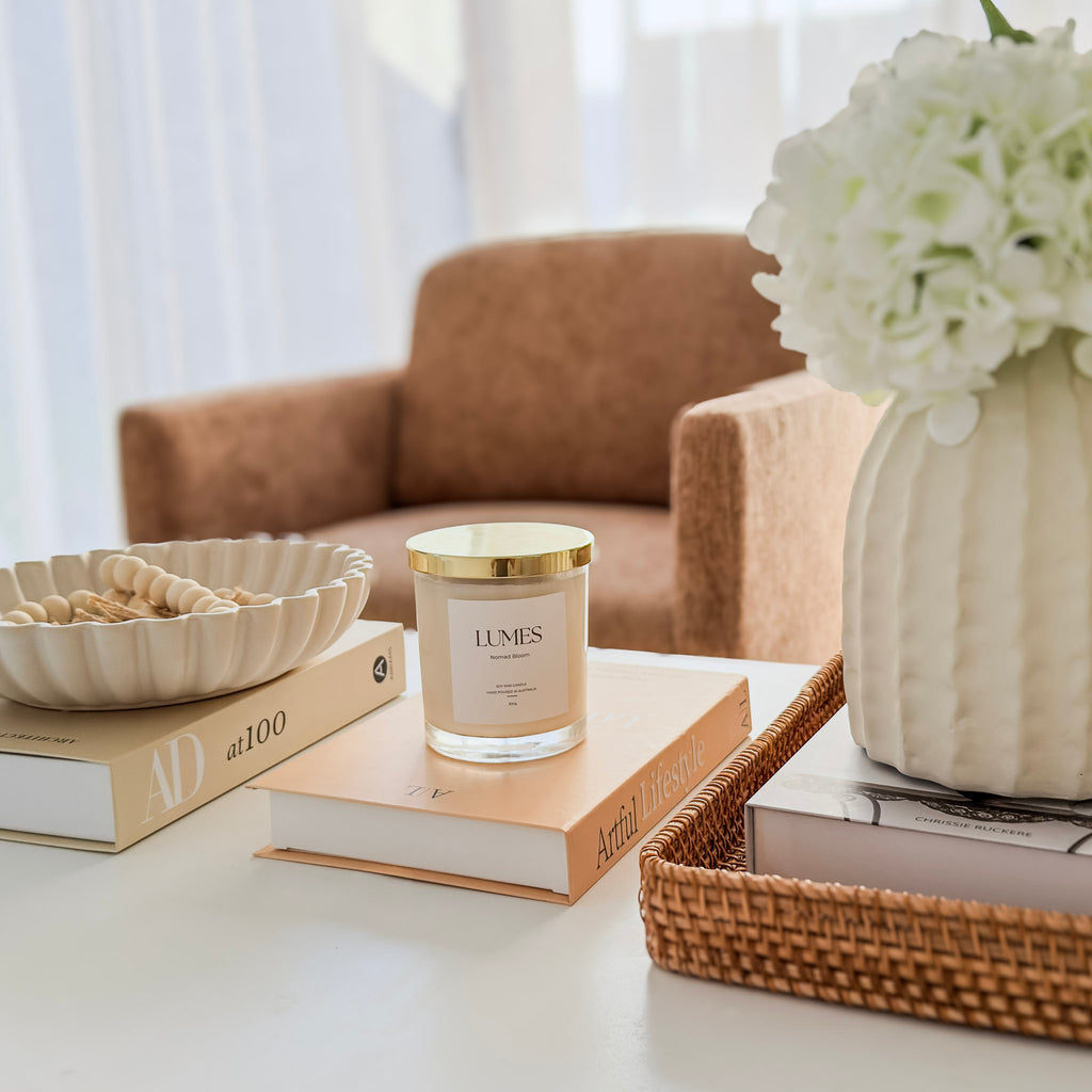 Candle with 'Lumes' branding on a table with books and a vase of flowers in a home setting.